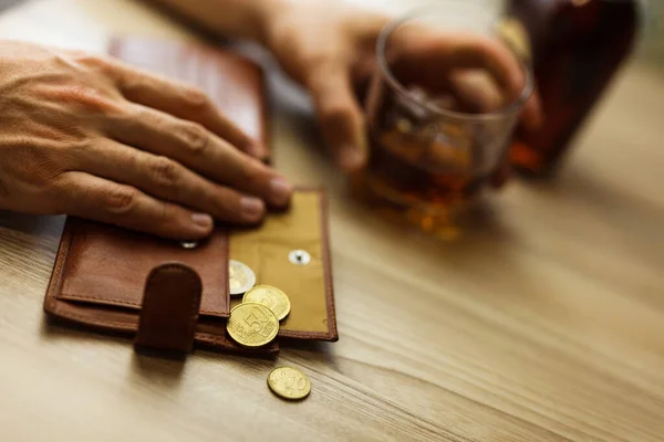 Fired man countsing his last money to drink expensive alcohol. Wrinkled alcoholic sits at brown wooden table with a glass of cognac with ice, on the table, empty wallet with coins
