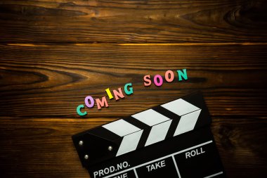 Coming soon Word made from multi-colored wooden letters on a brown wooden background and movie clapperboard. Filmmaking concept clipart