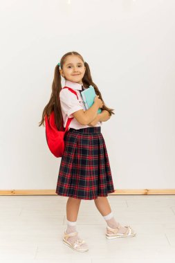 Sırt çantası beyaz arka planda duran ve elinde kitap tutan üniformalı bir kızın tam boy portresi.