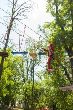 Parlak kırmızı eşofmanlı kız yüksek ipe tırmanıyor ve maceranın tadını çıkarıyor.