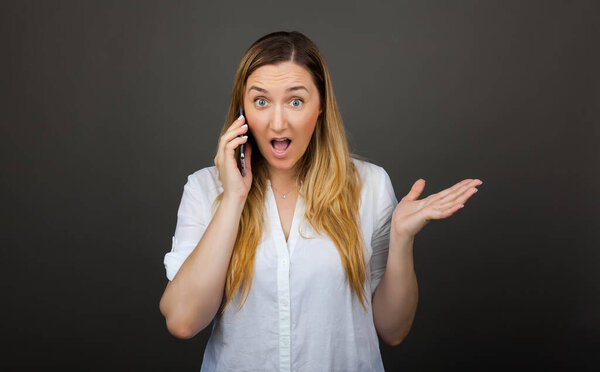 Surprised woman holding smartphone using mobile 30s aged talking on the phone amazedly looking at camera grey studio background