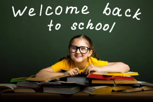 Welcome Back to school concept. Happy schoolgirl in glasses wearing a yellow t-shirt, at the green chalkboard in classroom. Chalk inscription Welcome Back to school.