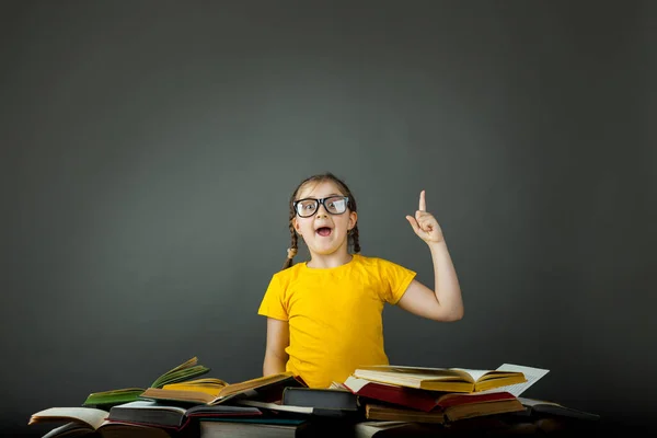 Back to school and happy time. Cute industrious child at desk indoors, desk pile of books. Child School Girl Pointing Blackboard, Kid Student over Black Board Background in Classroom