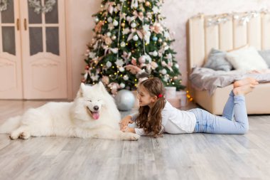 Gülümseyen köpeği Samoyed 'li Noel çocuğu kız büyük bir Noel ağacının önünde yere yattı. Noel, kış ve insanlar konsepti. Noel tebrik kartı. - Mutlu yıllar. Evde yeni yıl.