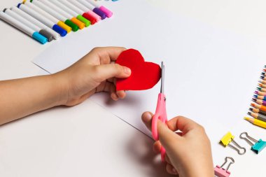 Çocuklar için kağıt işçiliği. Çocuk elleri masada makasla kağıttan kırmızı bir kalbi kesiyor. Çocuk sanatı. El işi konsepti. Bir tebrik kartı nasıl yapılır
