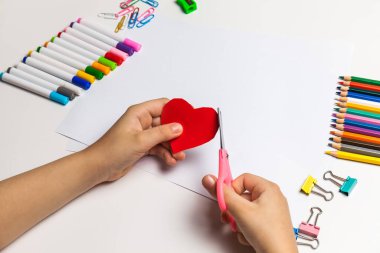 Çocuklar için kağıt işçiliği. Çocuk elleri masada makasla kağıttan kırmızı bir kalbi kesiyor. Çocuk sanatı. El işi konsepti. Bir tebrik kartı nasıl yapılır