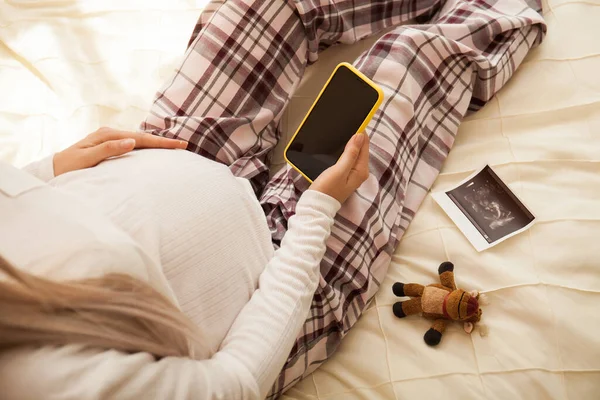 年轻的孕妇拿着智能手机躺在沙发上休息 床上的玩具和超声波扫描 — 图库照片