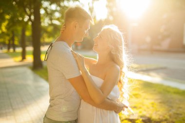Mutlu ve genç hamile bir çift, şehir parkında güneş ışığında sarılıyorlar. Yumuşak odak