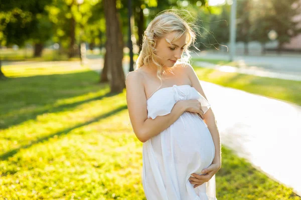 在阳光明媚的夏日 一位穿着白衣的快乐而自豪的孕妇站在公园里 背对着温暖的背光 看着自己的肚子 — 图库照片
