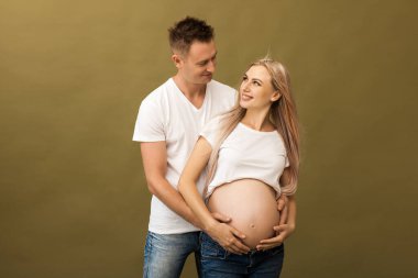 Bej arka planda gülümseyen hamile bir çift resmi. Koca, karısının karnına elleriyle dokunuyor.