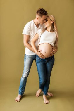 Bej arka planda gülümseyen bir çift resmi. Koca, karısının karnına elleriyle dokunuyor.