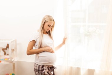 Genç, güzel, hamile bir kadın perdelerin ve pencerelerin yanında duruyor.