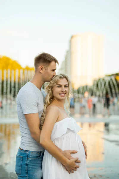 年轻漂亮的一对 身穿白衣的孕妇 在城市广场上 与喷泉拥抱在一起 — 图库照片