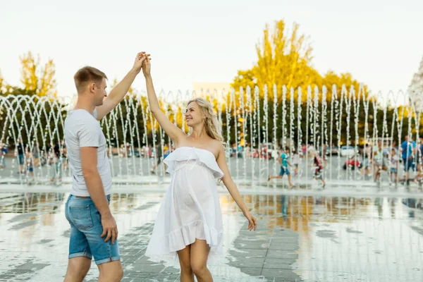 Young Caucasian couple in love walking and dancing on the city square with fountains. Pregnant woman in white dress dansing with her handsome man. Happy moments. and pregnancy concept.