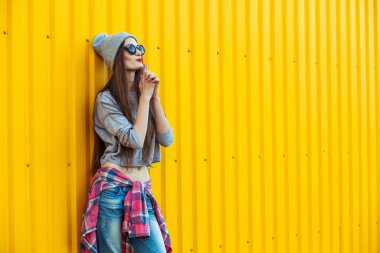 Güzel, uzun saçlı, şapkalı, güneş gözlüklü, parlak sarı arka planda poz veren genç bir kadın.