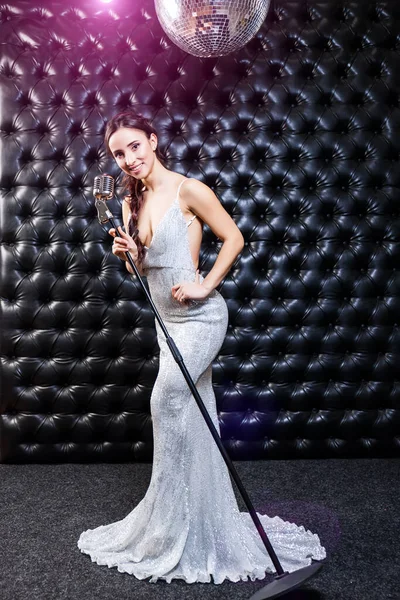 A beautiful young sexy woman in a long evening silver sparkling dress sings into a retro microphone in the dark. Black background.
