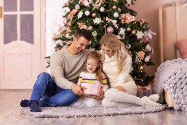 Üç kişilik aile yatak odasında büyük bir Noel ağacının önünde oturuyor, kız hediyesini açıyor. Noel hediyeleri konsepti