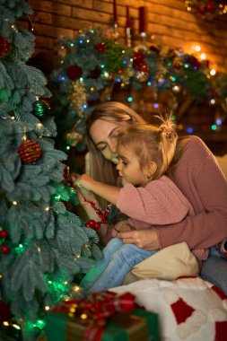 Sevgi dolu anne ve küçük kızı, Noel ağacını samimi ve şenlikli bir odada birlikte süsler. Sıcak ışıklarla dolu sahne, hassas bir aile tatili anını yakalıyor.