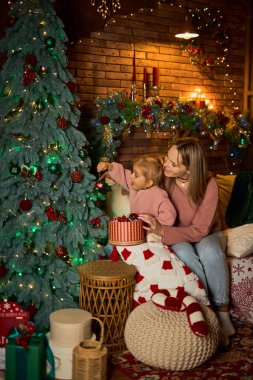 Sevgi dolu anne ve küçük kızı, Noel ağacını samimi ve şenlikli bir odada birlikte süsler. Sıcak ışıklarla dolu sahne, hassas bir aile tatili anını yakalıyor.