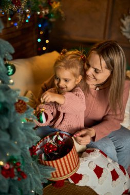 Sevgi dolu anne ve küçük kızı, Noel ağacını samimi ve şenlikli bir odada birlikte süsler. Sıcak ışıklarla dolu sahne, hassas bir aile tatili anını yakalıyor.