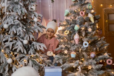 Şapkalı ve kazaklı bir çocuk iki süslü Noel ağacı arasında duruyor, hediye kutularına bakıyor. Fotoğraf bir tatil harikası ve beklenti hissi yakalıyor.