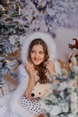 Beyaz kürk desenli pelerinli çekici kız, bir Noel stüdyosunda poz veriyor. Tatil promosyonları ve mevsimlik dekorlar için karlı ağaçların olduğu büyülü kış ortamı.