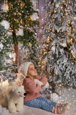 Süslü bir şapka ve kazak giyen çocuk, tuhaf bir ifadeyle karlı bir zeminde oturuyor, süslü Noel ağaçları ve kutup ayısı oyuncakları ile çevrili, büyülü bir kış sahnesini çağrıştırıyor.
