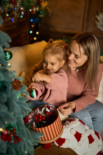 Sevgi dolu anne ve küçük kızı, Noel ağacını samimi ve şenlikli bir odada birlikte süsler. Sıcak ışıklarla dolu sahne, hassas bir aile tatili anını yakalıyor.