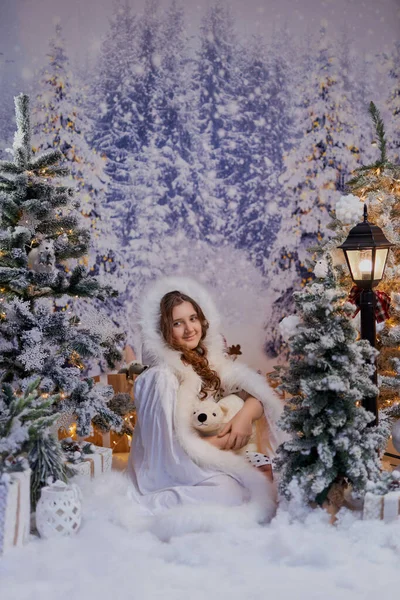 Beyaz kürk desenli pelerinli çekici kız, bir Noel stüdyosunda poz veriyor. Tatil promosyonları ve mevsimlik dekorlar için karlı ağaçların olduğu büyülü kış ortamı.