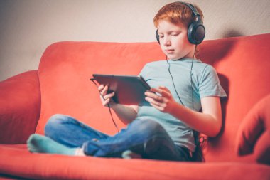 Bir tablette sesli kitap dinlerken kanepede oturan kızıl saçlı çocuk