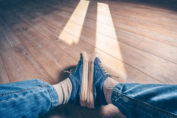 Kind from the first person on legs in jeans and sneakers sitting on a floor in beams of a sunlight