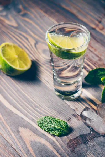 Cold vodka in a shot glass on a wooden table with a slice of lime, mint and ice slices