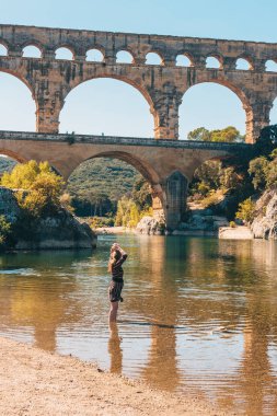 Vers-Pont-du-Gard, Gard / Occitanie / Fransa - 26 Eylül 2018: Avrupa görünümlü kız turist, antik Roma su kemeri Pont du Gard 'ın arka planında Gardon nehrinin kıyısında dinleniyor