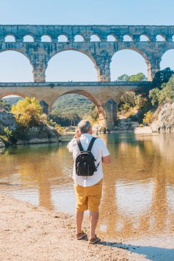 Vers-Pont-du-Gard, Gard / Occitanie / Fransa - 26 Eylül 2018: Avrupa kökenli bir erkek turist, Antik Roma su kemeri Pont du Gard'ın arka planına karşı Gardon nehri boyunca yürüyor