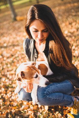 Siyah deri ceketli uzun saçlı esmer, evcil hayvanı Jack Russell Terrier ile parkta oynuyor.