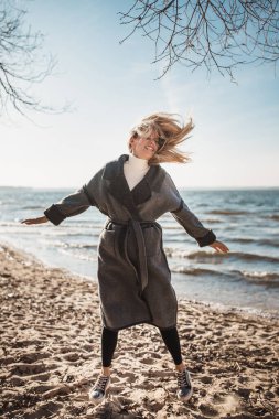 Uçarı, büyüleyici, karizmatik sarışın rüzgarlı bir günde kumlu sahilde neşeyle zıplıyor.
