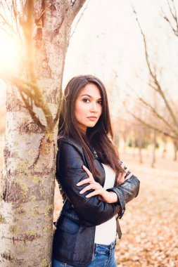 Ciddi odaklı esmer kız ellerini göğsüne koydu. Parktaki bir ağaca yaslandı. İş portresi.