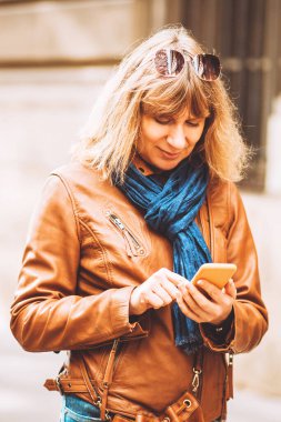 Pahalı deri ceketli güzel bir kadın ve akıllı telefonlu bir şehir caddesinde günlük giysiler. Bilgiye bakar ve mali durumu kontrol eder.