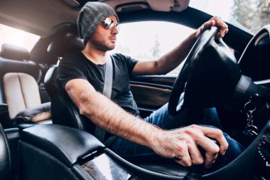 Güçlü bir arabanın içinde, şapkalı ve güneş gözlüklü bir adam güvenli bir şekilde direksiyonun arkasında manüel mekanik şanzıman kullanıyor.