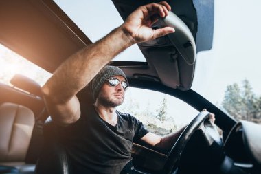 Kafkasyalı gözlüklü ve şapkalı bir adam yolcu kompartımanında direksiyon başında otururken salonun dikiz aynasını ayarlıyor.