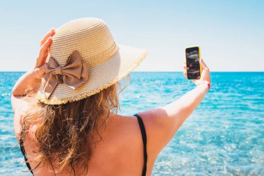 Şapkalı güzel sarışın kadın denizin arka planında özçekim yapıyor. Tropik sahillerde tatil sezonu.