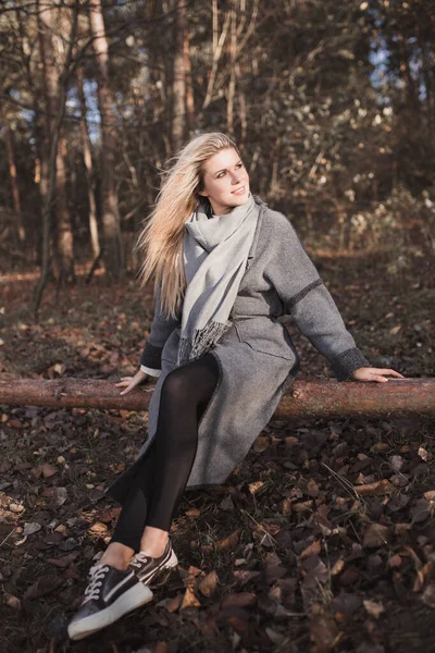 Güzel bir sarışının sonbahar portresi, sıcacık bir ceketle ormanda bir kütüğün üzerinde güneşlenirken.