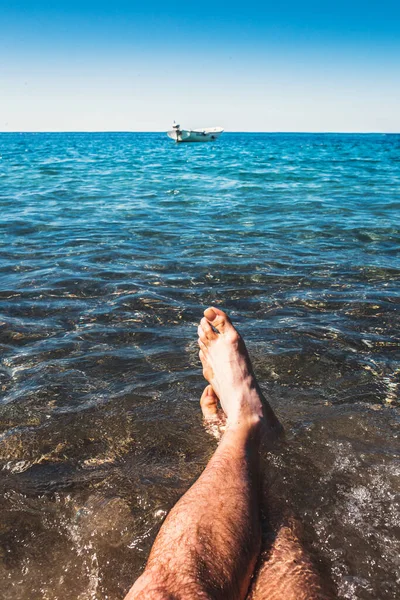 Male feet on the seashore - near the shore a boat on the horizon - uninhabited island