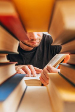Kitap yığınlarının arasından kitap okuyan bir adam. Okumaya hevesli.