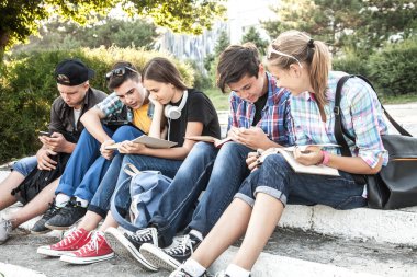 Kitapları ve aletleri olan bir grup genç öğrenci parktaki merdivenlere oturur.