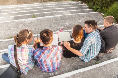 Kitapları ve aletleri olan bir grup genç öğrenci parktaki merdivenlere oturur.