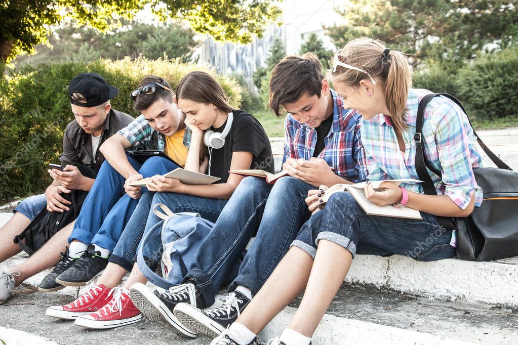 group of young students with books and gadgets sit on the steps in the park