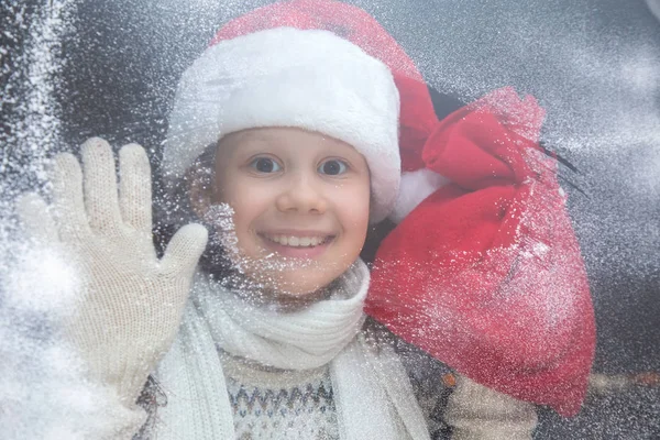 冬のストリートから雪に覆われたウィンドウを調べてプレゼントとサンタの帽子の少女 — ストック写真