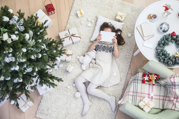 少女はカーペット上でタブレットとクリスマス ツリーの下に横たわっています 上からの眺め — ストック写真