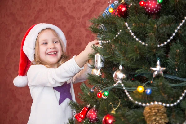 サンタの帽子の女の子はクリスマスツリーを飾る — ストック写真
